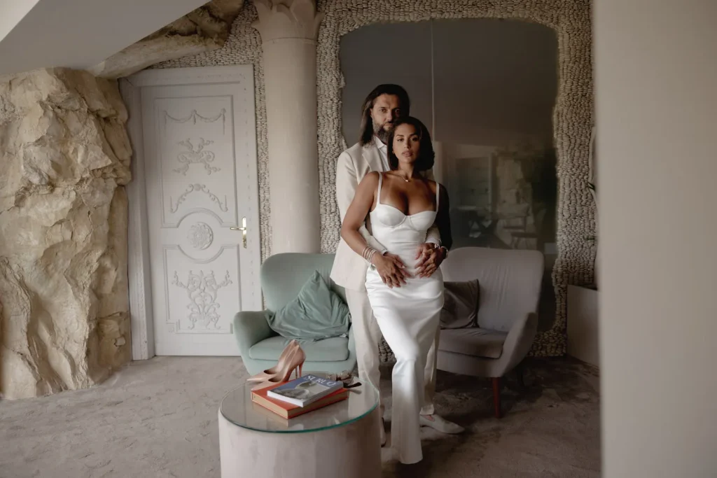 Luxury Couple Shoot in Taormina | Italian Glamour 33 A couple poses indoors; the woman wears a white dress and the man wears a light suit. They stand in front of a chair and small table with shoes and books, set against textured walls and an ornate door.