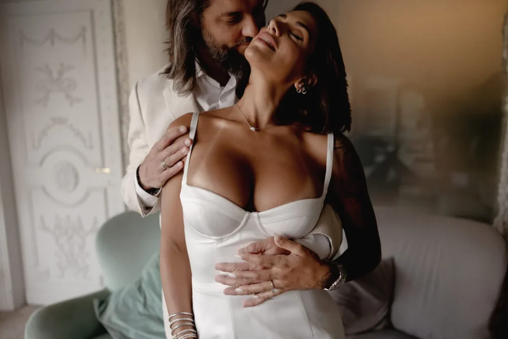 Luxury Couple Shoot in Taormina | Italian Glamour 12 A man gently hugs and kisses a smiling woman dressed in a white dress. They appear happy and intimate, standing together in a softly lit, elegant room with ornate decor.