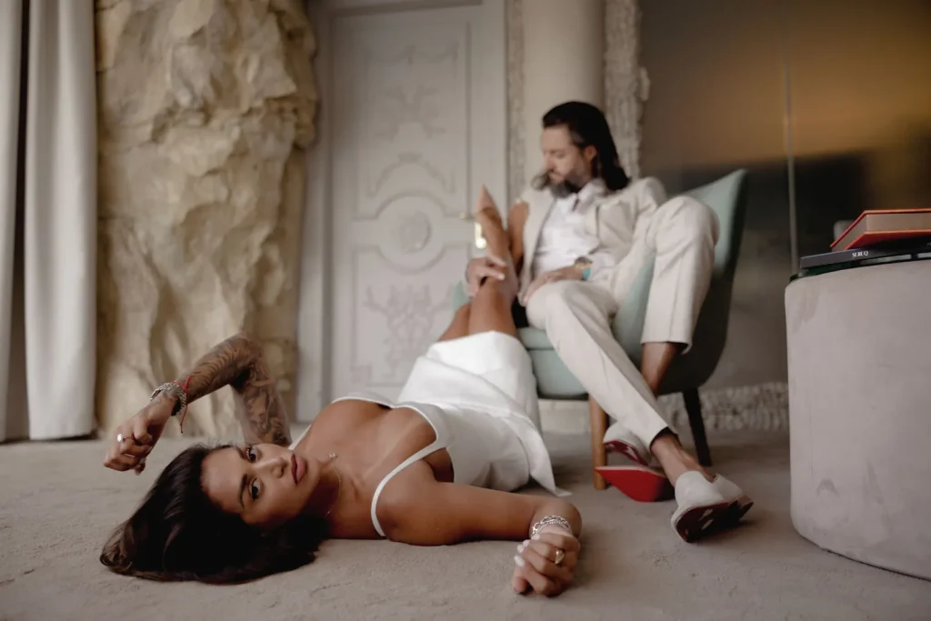 Luxury Couple Shoot in Taormina | Italian Glamour 39 A woman in a white dress lies on the floor, looking at the camera, while a man in a light suit sits behind her in a chair, holding her ankle. The setting appears luxurious and stylish.