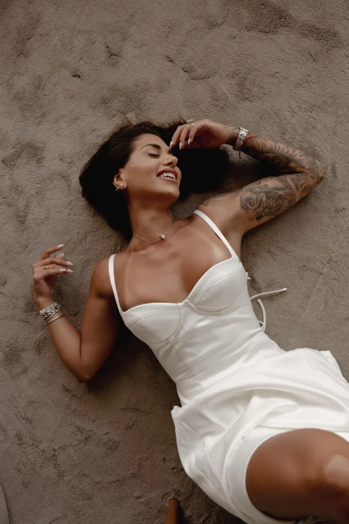 Luxury Couple Shoot in Taormina | Italian Glamour 35 A woman in a white dress lies on a beige surface, smiling with her eyes closed. She has tattoos on her left arm and is wearing jewelry, including bracelets, a necklace, and a watch.