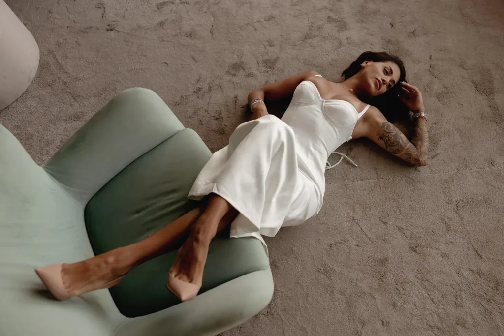 Luxury Couple Shoot in Taormina | Italian Glamour 48 A woman in a white dress and nude heels lies on a carpeted floor next to a green armchair, looking relaxed with one arm resting above her head.