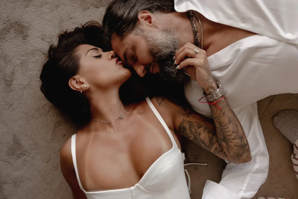 Luxury Couple Shoot in Taormina | Italian Glamour 36 A woman in a white dress and a man in a white shirt lie on the floor, facing each other closely. The woman kisses the man on his forehead. They both appear relaxed and affectionate.