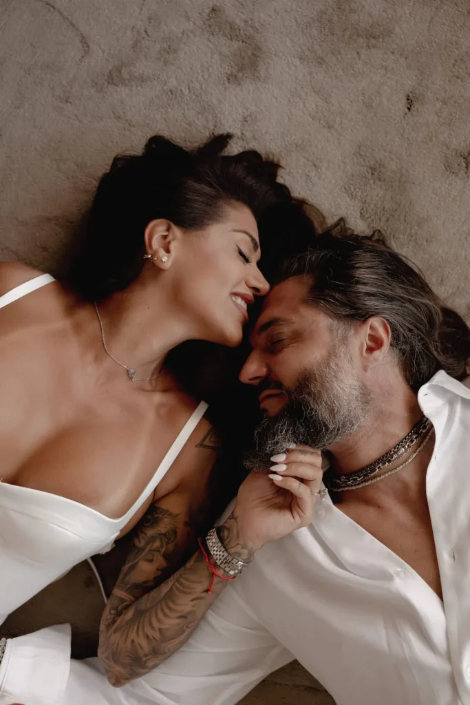 Luxury Couple Shoot in Taormina | Italian Glamour 5 A woman and man lie close together on a neutral surface, facing each other and smiling affectionately. The woman wears a white outfit, and the man wears a white shirt with jewelry. They appear relaxed and intimate.