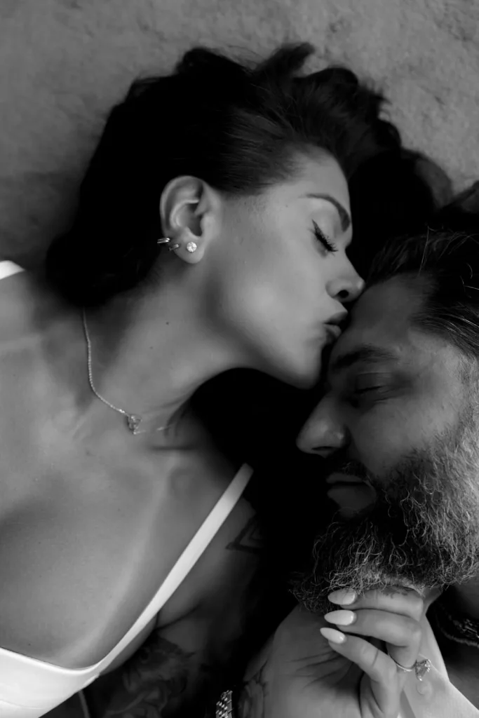 Luxury Couple Shoot in Taormina | Italian Glamour 2 A woman kisses a bearded mans forehead as they lie close together on a textured surface, both with eyes closed, in a black and white photo.