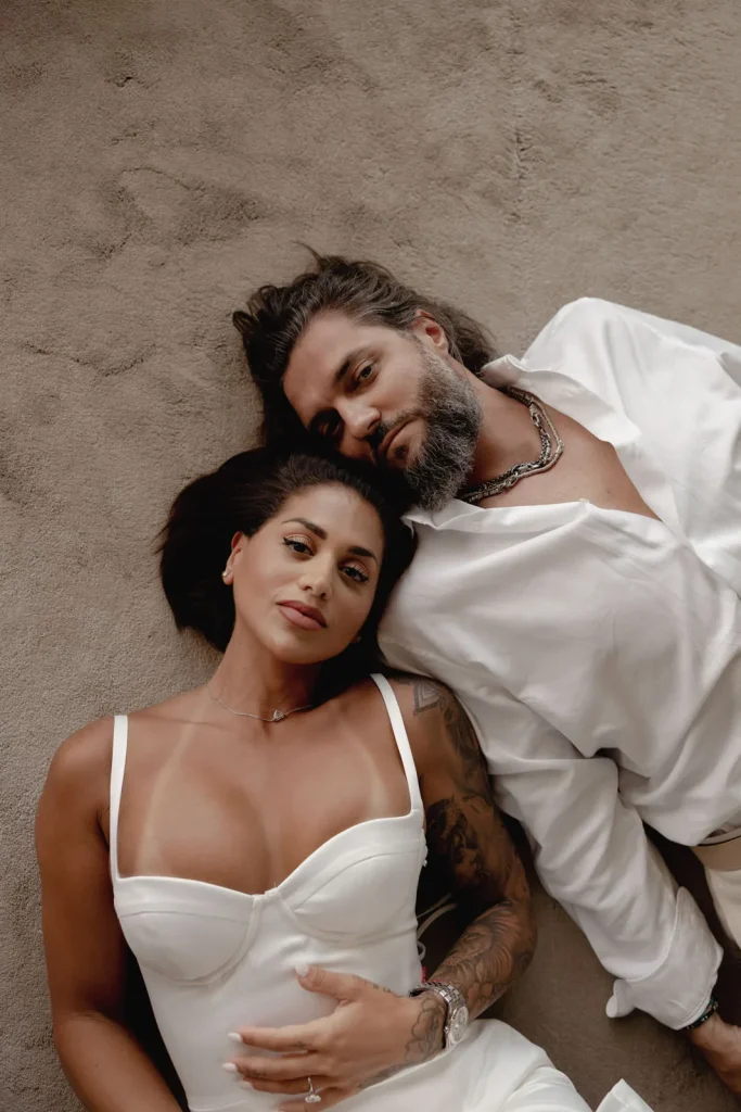 Luxury Couple Shoot in Taormina | Italian Glamour 69 A woman in a white dress and a man in a white shirt lie side by side on a beige surface, gazing calmly at the camera.