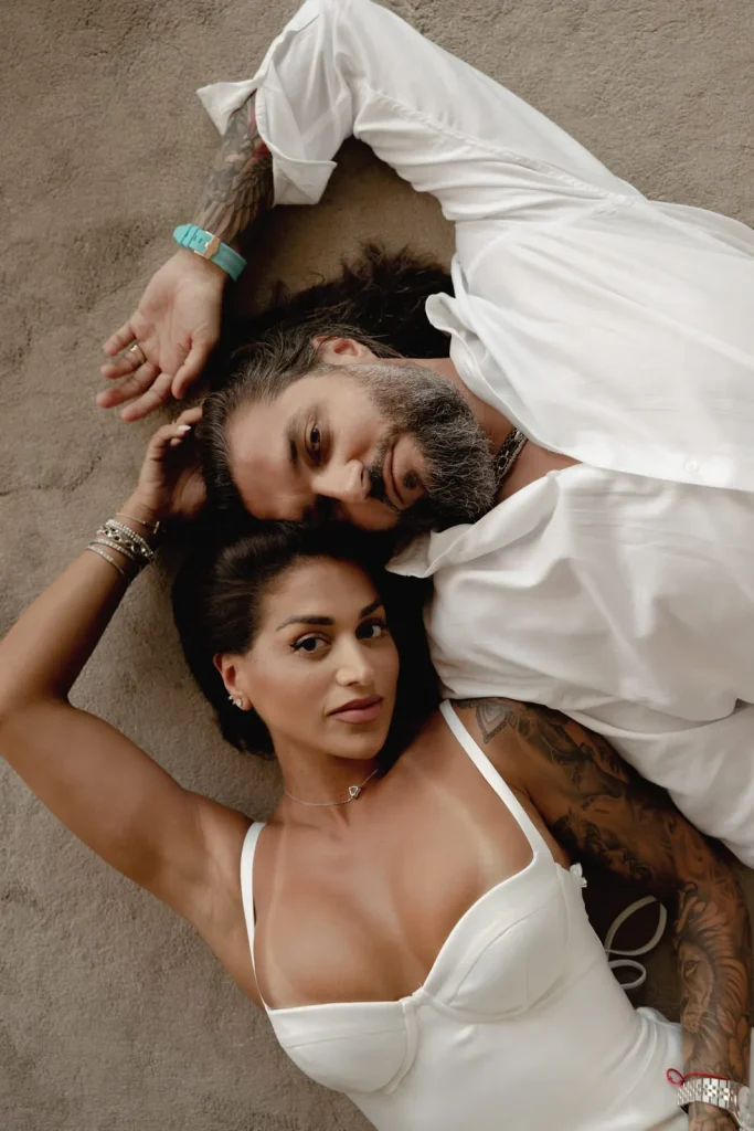 Luxury Couple Shoot in Taormina | Italian Glamour 14 A man and woman lie on a light-colored surface, heads touching. Both wear white clothing and have tattoos. The woman looks at the camera while the man gazes upward, their arms intertwined above their heads.