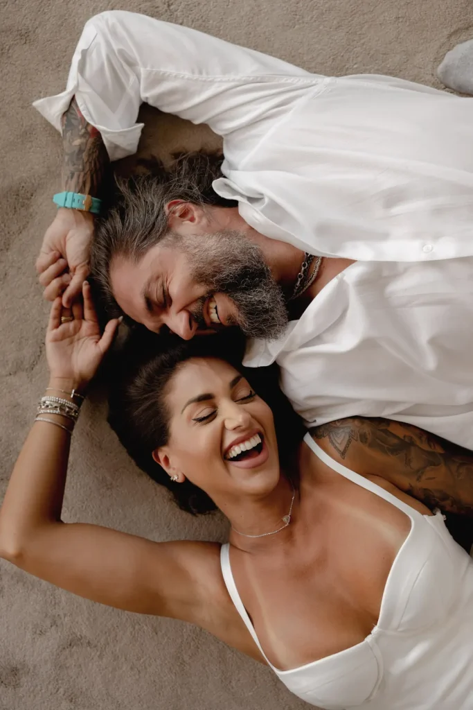 Luxury Couple Shoot in Taormina | Italian Glamour 68 A smiling couple lies on a beige carpet. The man has a beard, gray hair, tattoos, and wears a white shirt. The woman has dark hair, tattoos, a white top, and jewelry. Both look relaxed and happy.