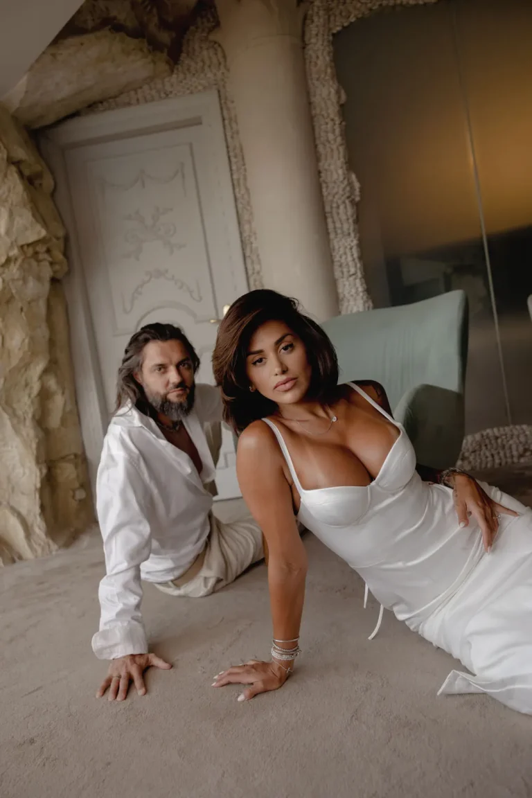 A woman in a white dress and a man in a white shirt sit on the floor indoors near a decorative wall, both looking toward the camera with relaxed expressions.