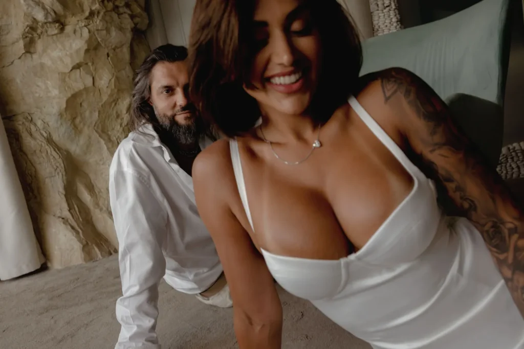 Luxury Couple Shoot in Taormina | Italian Glamour 34 A smiling woman in a white dress poses in the foreground, while a man with long hair and a beard, wearing a white shirt, sits in the background indoors.
