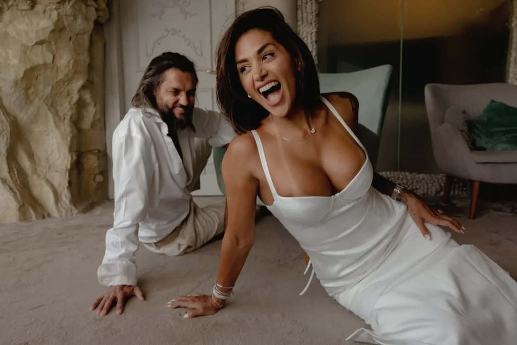 Luxury Couple Shoot in Taormina | Italian Glamour 61 A woman in a white dress laughs while sitting on the floor, with a smiling man in a white shirt sitting behind her. They appear relaxed and happy in a cozy, warmly lit room.