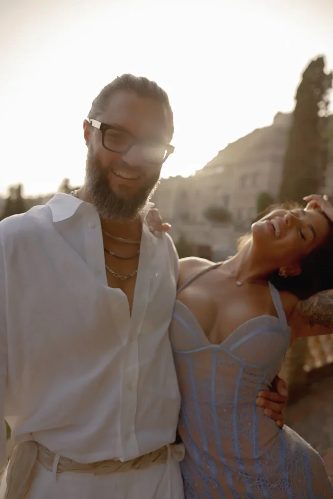 Luxury Couple Shoot in Taormina | Italian Glamour 56 A bearded man in glasses and a white shirt stands beside a smiling woman in a light blue lace dress, both enjoying sunlight outdoors with trees and buildings in the background.