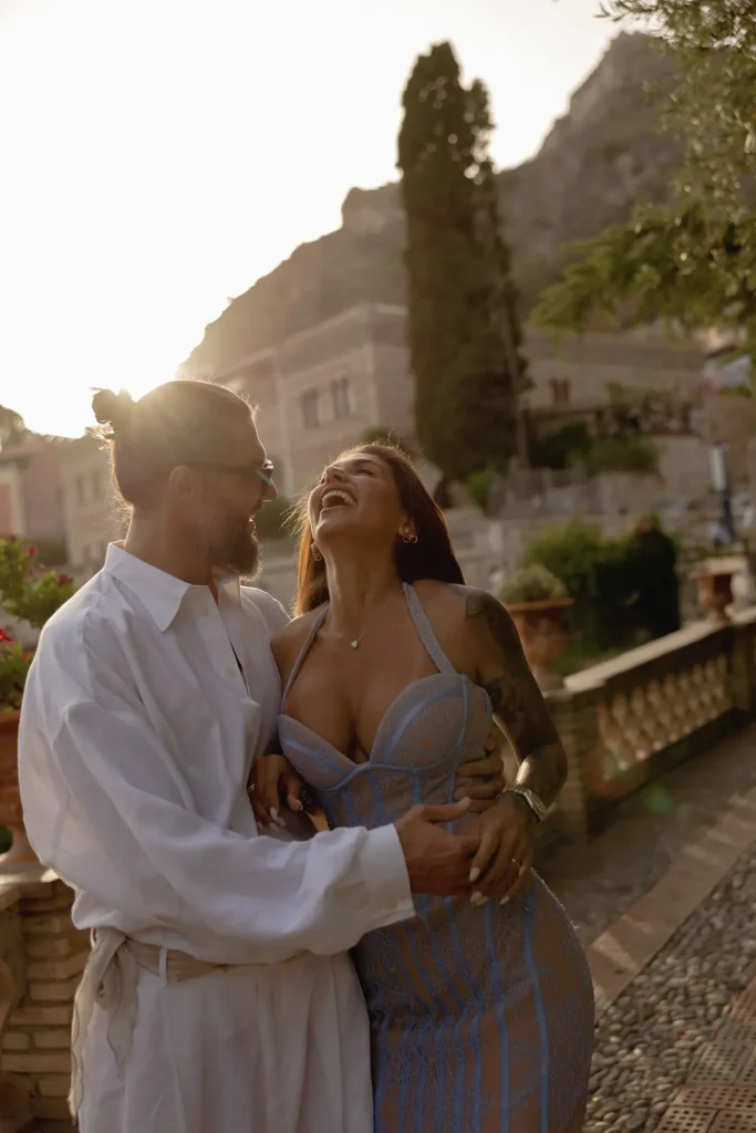 Luxury Couple Shoot in Taormina | Italian Glamour 22 A smiling couple stands outdoors in warm sunlight, the man in a white shirt and glasses holding a woman in a blue dress who is laughing, with scenic buildings and trees in the background.