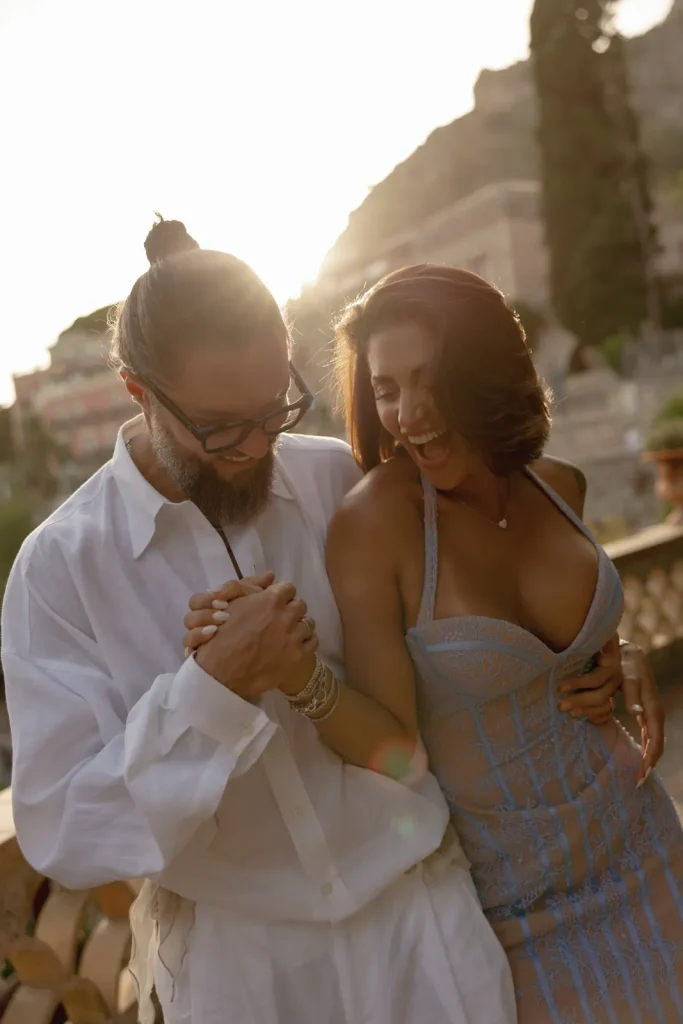 Luxury Couple Shoot in Taormina | Italian Glamour 59 A man in glasses and a white shirt smiles while holding hands with a woman in a light blue dress. They are outdoors at sunset, both laughing and enjoying each others company.