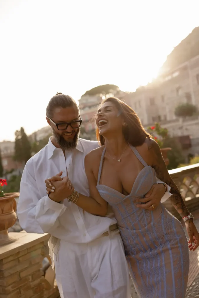 Luxury Couple Shoot in Taormina | Italian Glamour 1 A smiling couple walks arm in arm outdoors at sunset. The man wears glasses and a white shirt, while the woman wears a light blue dress. They appear happy and are laughing together.