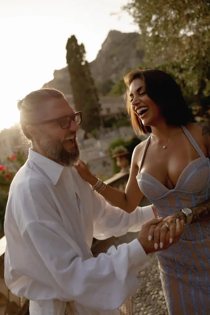 Luxury Couple Shoot in Taormina | Italian Glamour 26 A smiling man and woman hold hands and laugh outdoors at sunset. The man wears glasses and a white shirt; the woman wears a lacy dress. Trees and a building are visible in the background.