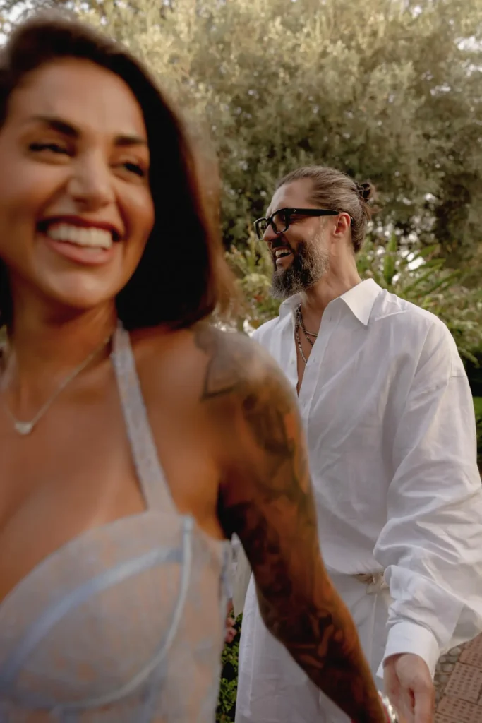 Luxury Couple Shoot in Taormina | Italian Glamour 32 A smiling woman in a light dress leads a bearded man in glasses and a white shirt outdoors. Both appear happy, with greenery and trees in the background.