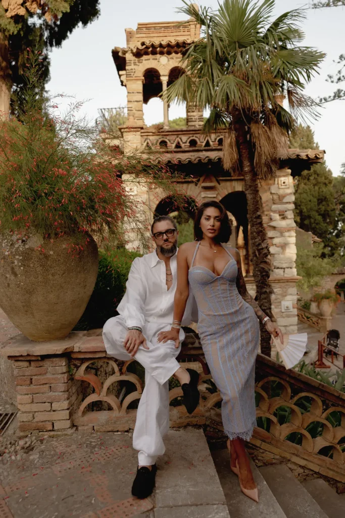 Luxury Couple Shoot in Taormina | Italian Glamour 7 A man in white clothes and a woman in a fitted dress pose by a stone railing in a lush garden with palm trees and an ornate, rustic building in the background.