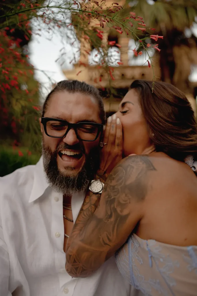 Luxury Couple Shoot in Taormina | Italian Glamour 65 A woman with tattoos whispers into a bearded mans ear, making him laugh. They are outdoors among greenery and red flowers, both appear joyful and close.