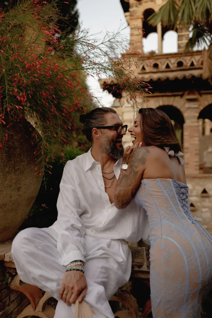 Luxury Couple Shoot in Taormina | Italian Glamour 10 A man in a white outfit and a woman in a light blue dress smile and sit closely together outdoors near a large vase and rustic architecture.