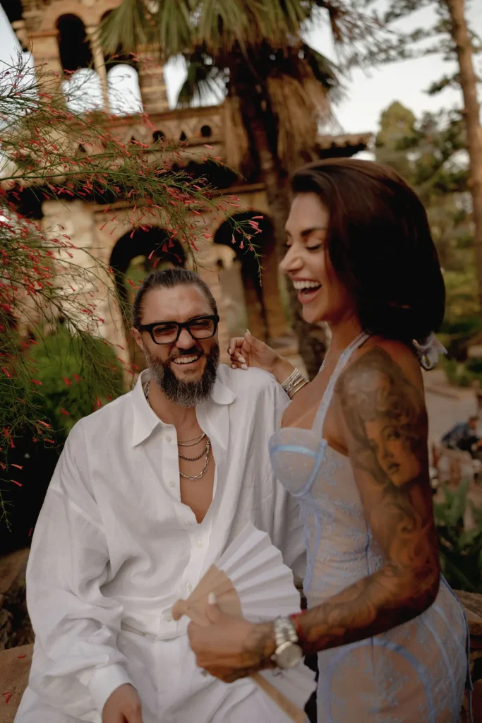 Luxury Couple Shoot in Taormina | Italian Glamour 13 A smiling man in a white shirt sits next to a laughing woman in a light blue dress with tattoos, outdoors near green plants and red flowers.