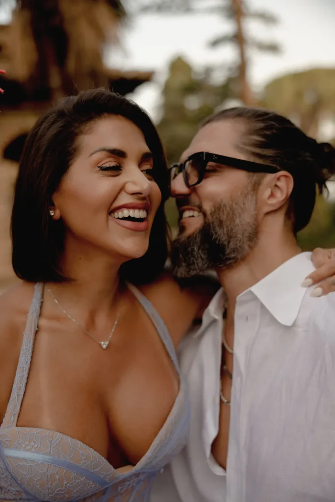 Luxury Couple Shoot in Taormina | Italian Glamour 6 A smiling woman in a blue dress stands close to a bearded man in glasses and a white shirt. They look happy and affectionate outdoors, with trees blurred in the background.