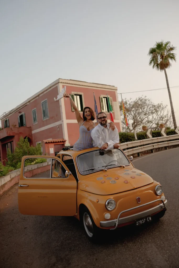 Luxury Couple Shoot in Taormina | Italian Glamour 47 A smiling couple poses with an open yellow vintage Fiat 500. The woman sits on the door waving papers, and the man stands inside. A pink building, palm tree, and flags are visible in the background on a sunny day.