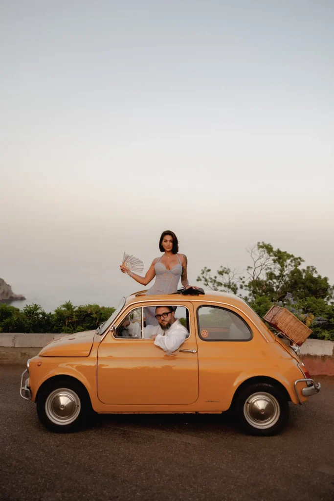 Luxury Couple Shoot in Taormina | Italian Glamour 51 A woman stands through the sunroof of a vintage yellow car, holding a fan. A man sits inside, wearing sunglasses and hat. Trees and a basket are visible in the background.