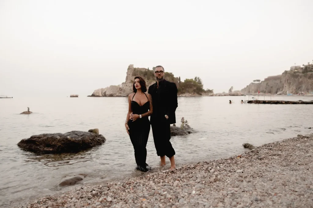 Luxury Couple Shoot in Taormina | Italian Glamour 62 A woman and a man in dark clothing stand barefoot on a rocky beach, with calm water, rocks, and a distant cliff in the background under a clear sky.