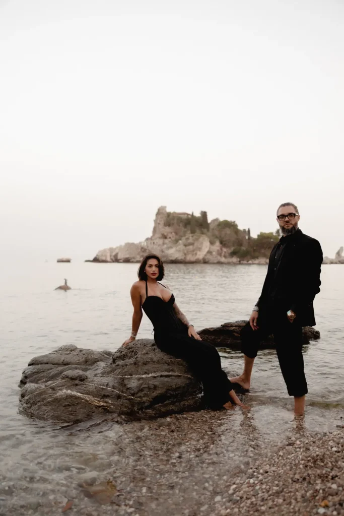 Luxury Couple Shoot in Taormina | Italian Glamour 40 A woman in a black dress sits on a rock, and a man in black stands barefoot beside her in shallow water. An island is visible in the background under a pale sky.