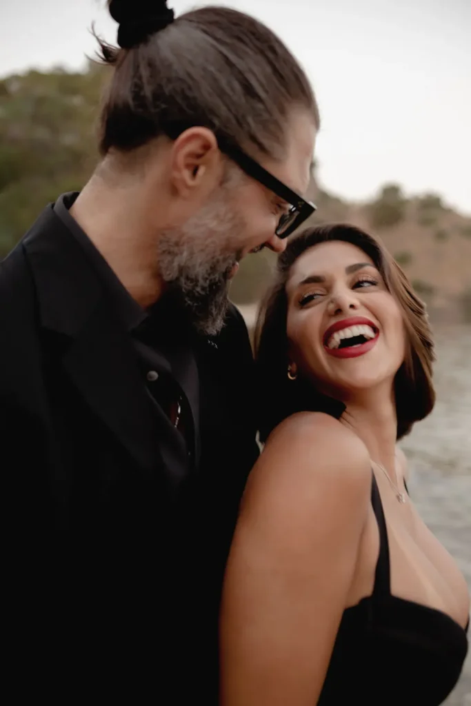 Luxury Couple Shoot in Taormina | Italian Glamour 28 A man and woman smile and laugh together outdoors, dressed in black. The woman looks up at the man with a big smile, and they appear happy and close. Trees and water are blurred in the background.