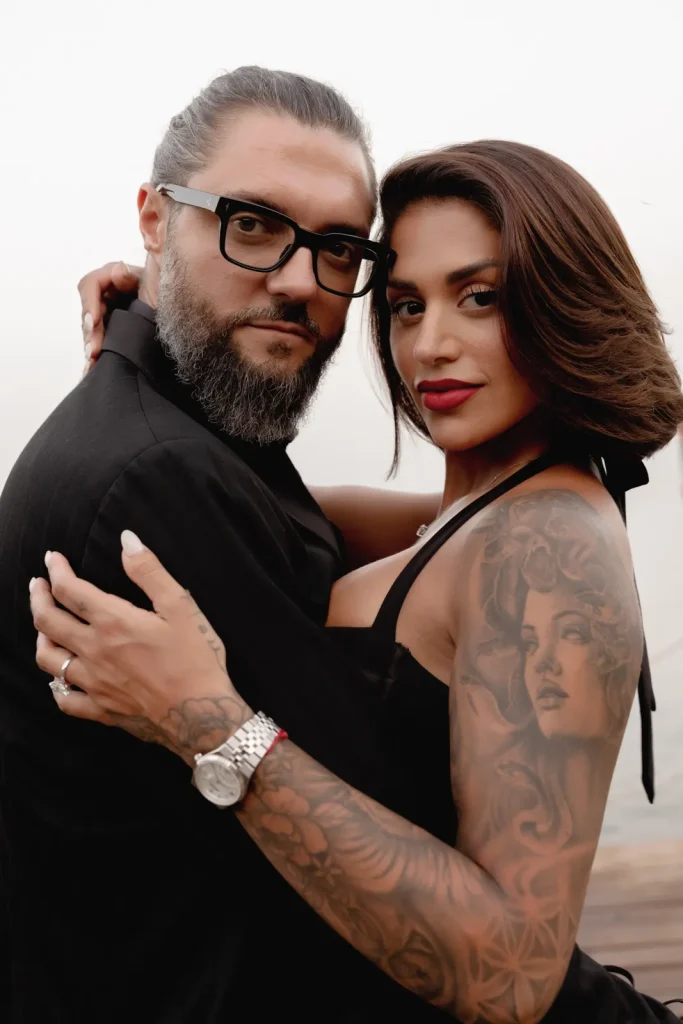 Luxury Couple Shoot in Taormina | Italian Glamour 24 A man with glasses and a beard embraces a woman with long hair, bold makeup, and detailed tattoos on her arm. They both wear black and look confidently at the camera against a blurred light background.