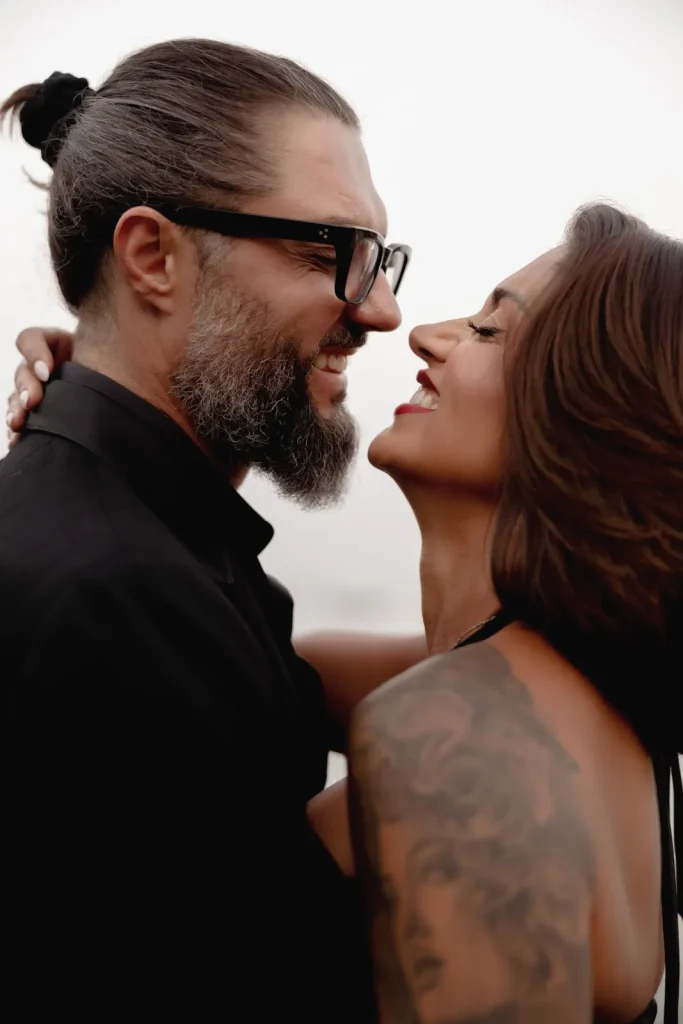 Luxury Couple Shoot in Taormina | Italian Glamour 4 A smiling couple faces each other closely. The man has a beard, glasses, and a man bun. The woman has long hair, red lipstick, and a tattoo on her arm. They appear happy and affectionate.