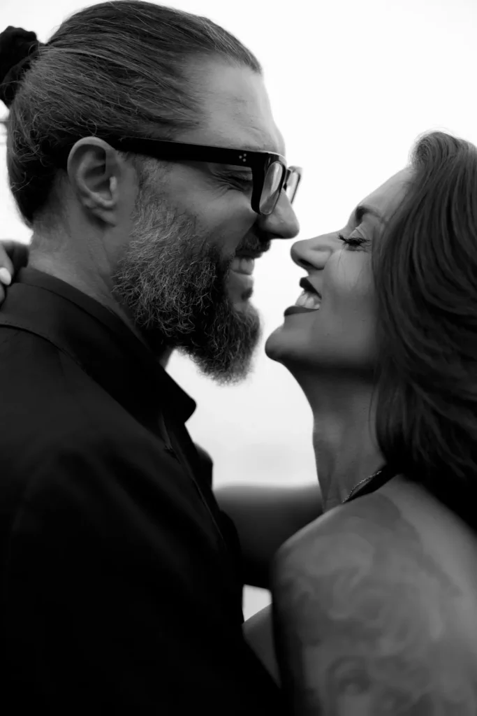 Luxury Couple Shoot in Taormina | Italian Glamour 20 A bearded man with glasses and a woman with long hair smiling at each other closely, sharing an affectionate moment. The photo is in black and white.