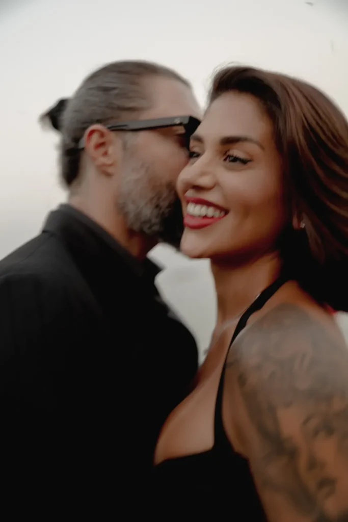 Luxury Couple Shoot in Taormina | Italian Glamour 54 A man with a beard whispers into a smiling womans ear. The woman has a tattoo on her arm and bright red lipstick. Both appear happy and close together.