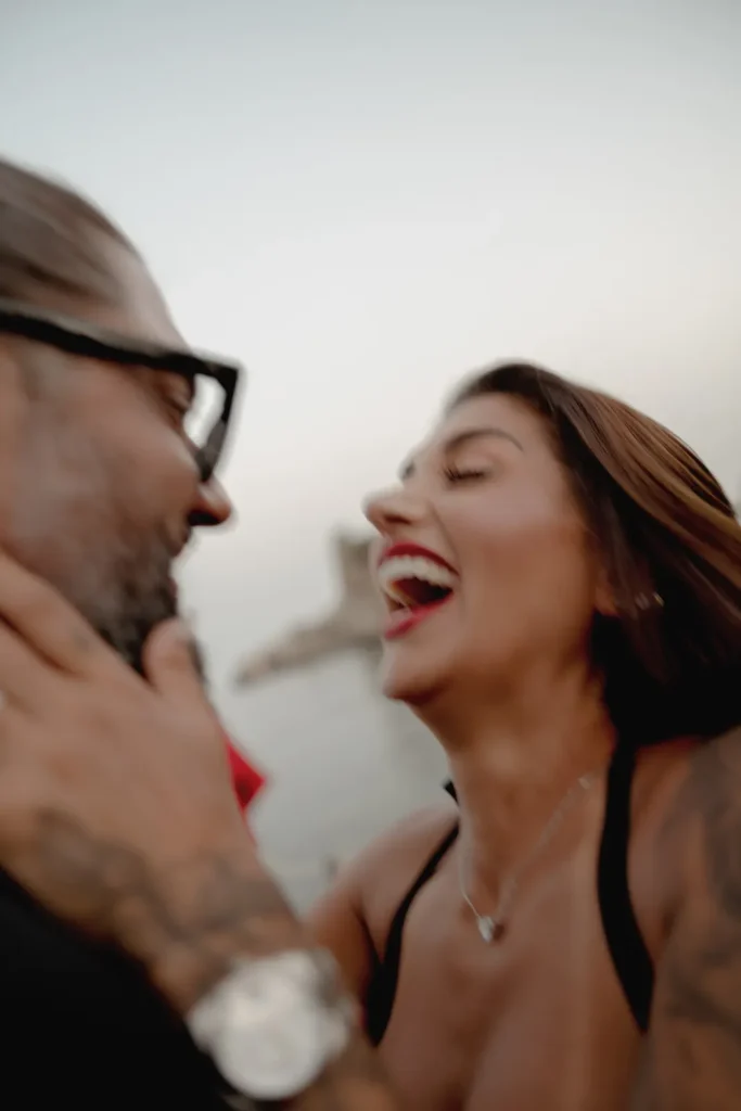 Luxury Couple Shoot in Taormina | Italian Glamour 50 A man and woman laugh together outdoors, faces close and joyful. The image is slightly blurred, capturing a candid, happy moment between them.