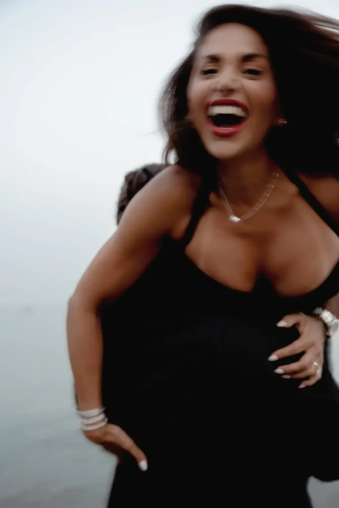 Luxury Couple Shoot in Taormina | Italian Glamour 37 A woman in a black dress smiles and laughs, being lifted up by someone partially visible. She has long dark hair and is wearing jewelry, with a blurred outdoor background.