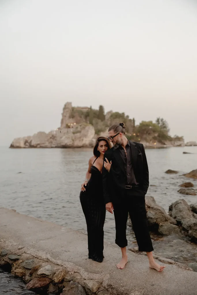 Luxury Couple Shoot in Taormina | Italian Glamour 27 A man and woman in formal black outfits stand on a rocky shoreline at sunset, with an island and calm water in the background. The man is barefoot and wears sunglasses.