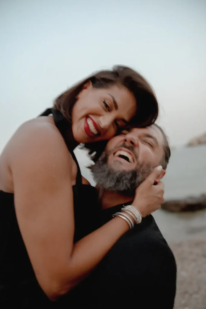 Luxury Couple Shoot in Taormina | Italian Glamour 17 A woman in a black dress hugs a bearded man and they both laugh joyfully on a beach, with water and rocks in the background.